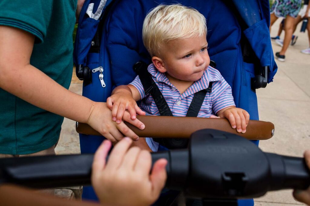A toddler is strapped into a bright blue stroller. He holds onto a boys hand while another boys hand is in the foreground holding onto the stroller handle. 
