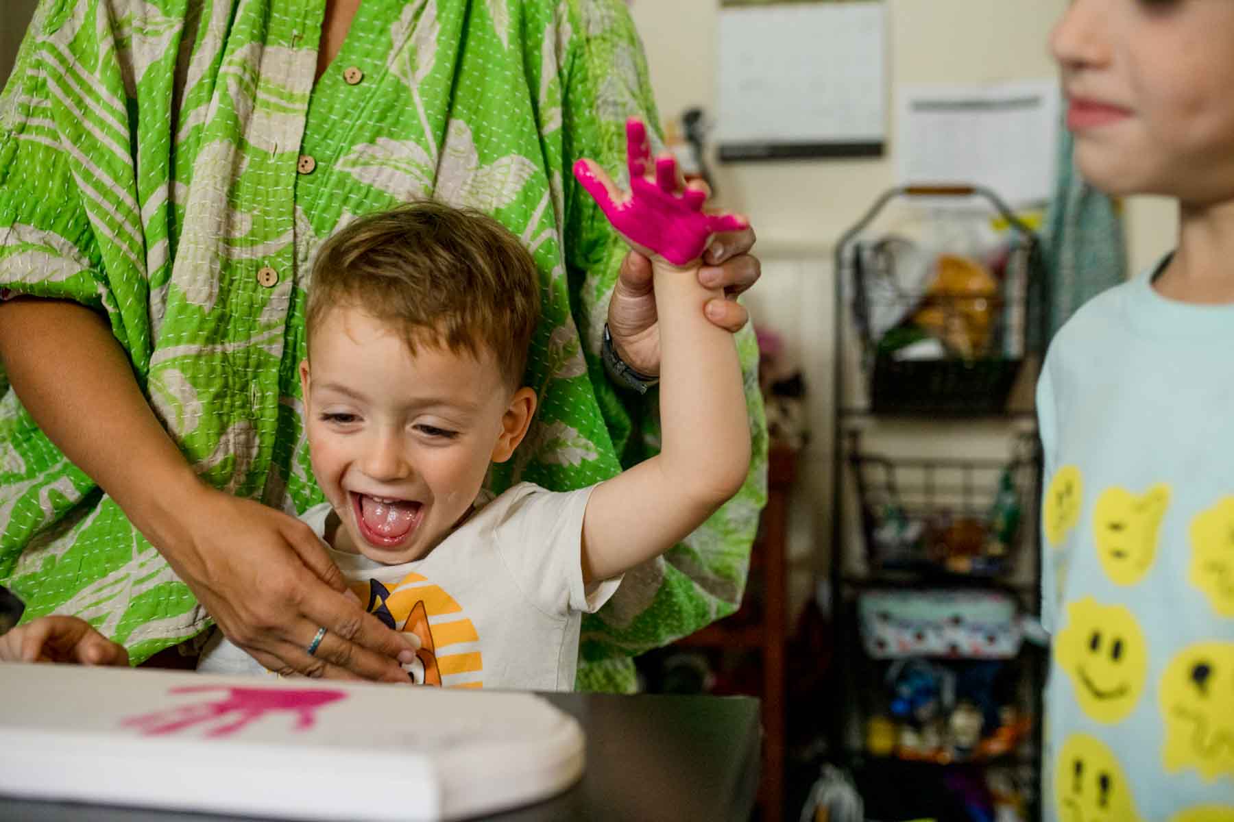 A little boy wearing a white shirt sits at a table with his hand covered in bright pink paint with his moms hand holding his over his head.