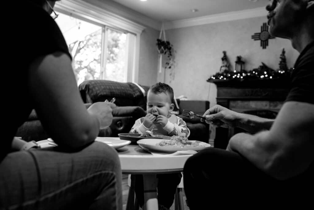 Mom and dad in the foreground sitting at their toddler's table eating dinner with him. The toddler has his hands in front of his mouth in the center of the image. 