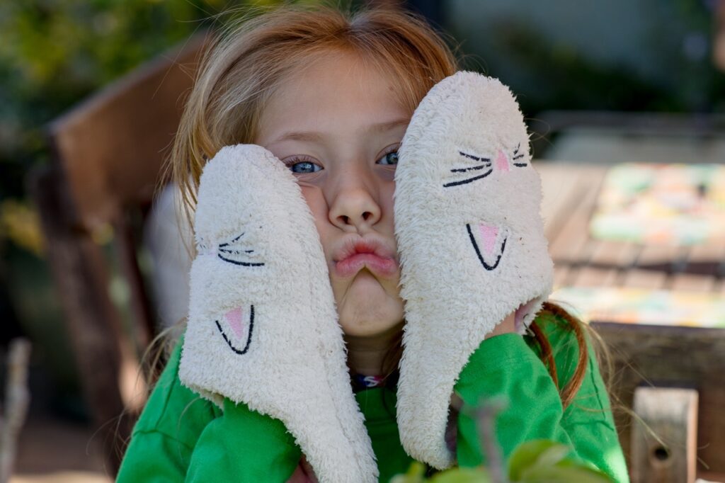 A young girl with blue eyes smushes her cheeks and makes a pouty face with her white and pink bunny slippers on her hands. 