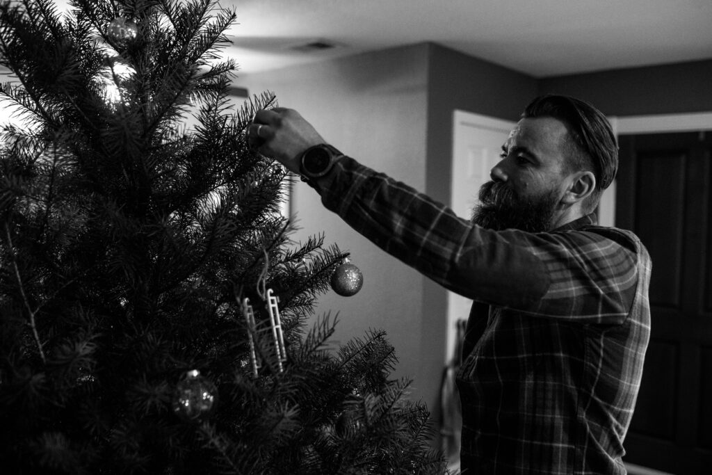 A man wearing a plaid shirt reaches up to hang an ornament on the Christmas tree. 