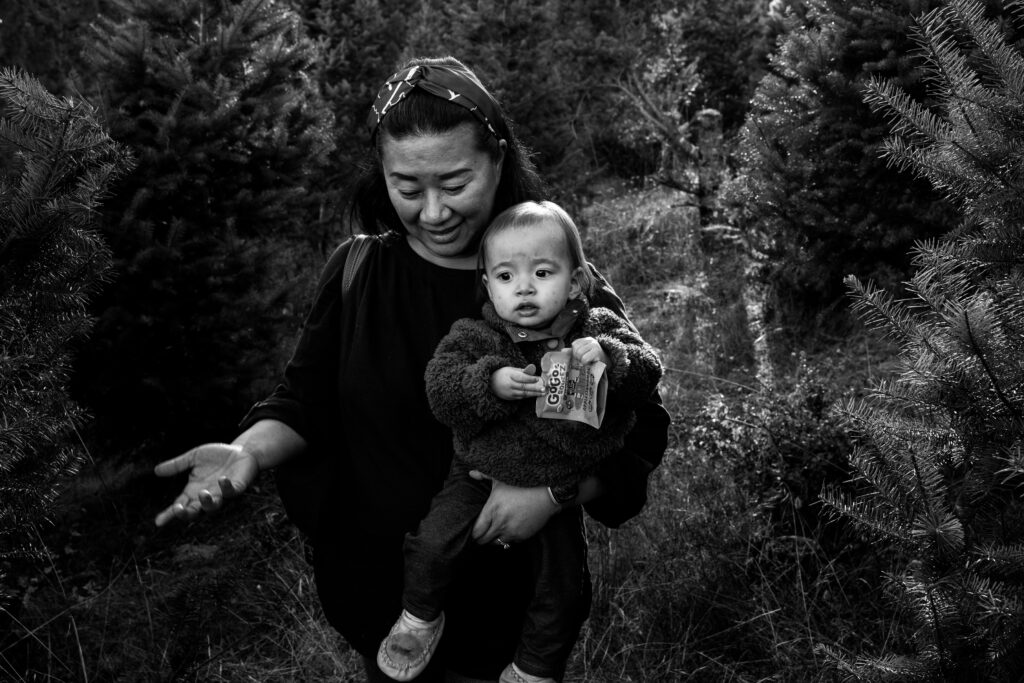 A mom carrying a small toddler gestures in a quizzical way while walking between fir trees. 