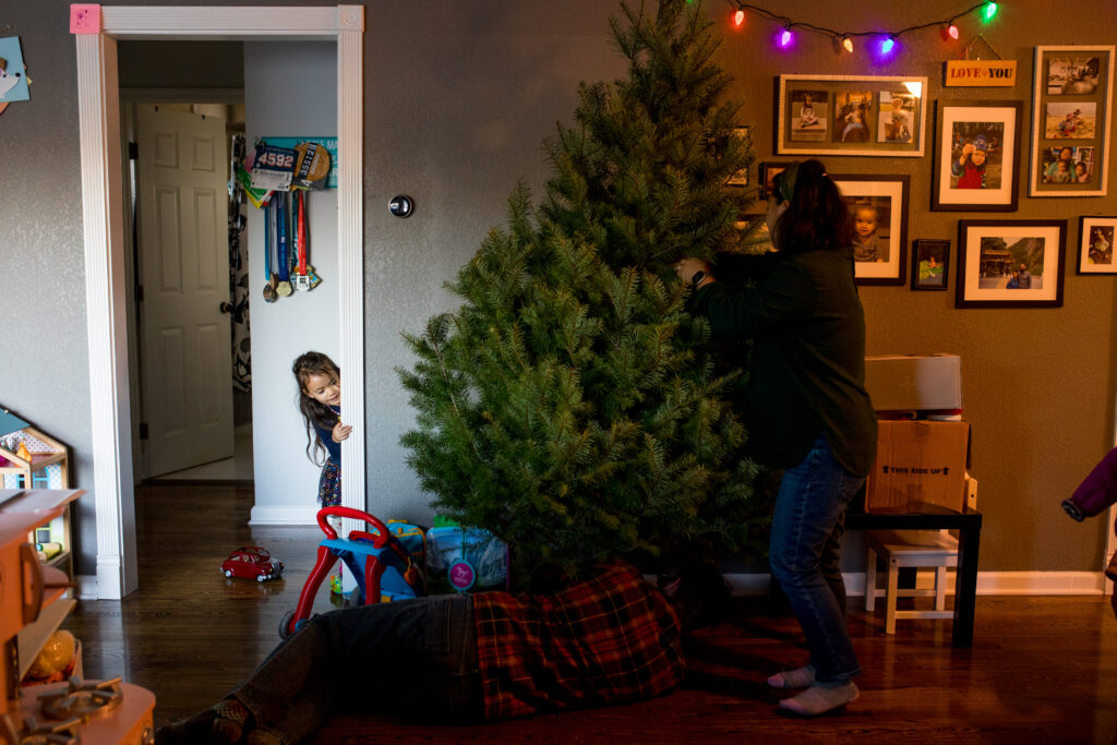 Mom stands and holds Christmas tree in the stand as dad lies on the ground securing it, while their daughter peeks around the corner at them.