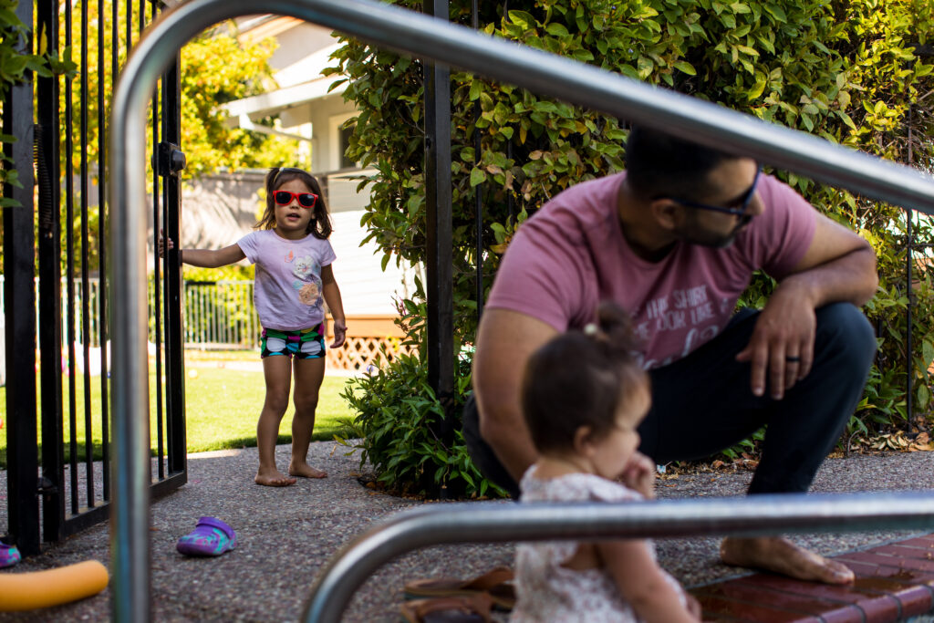 A little girl in a bathing suit and wearing bright red sunglasses opens the gate to the pool where her dad and baby sister sit at the pools edge, framed by the pool railing. 