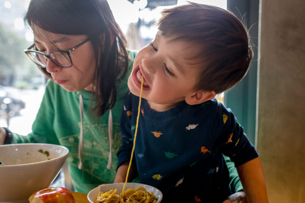 A mom holds her toddler son who is wearing a dinosaur print shirt as he slurps noodles at a restaurant. 