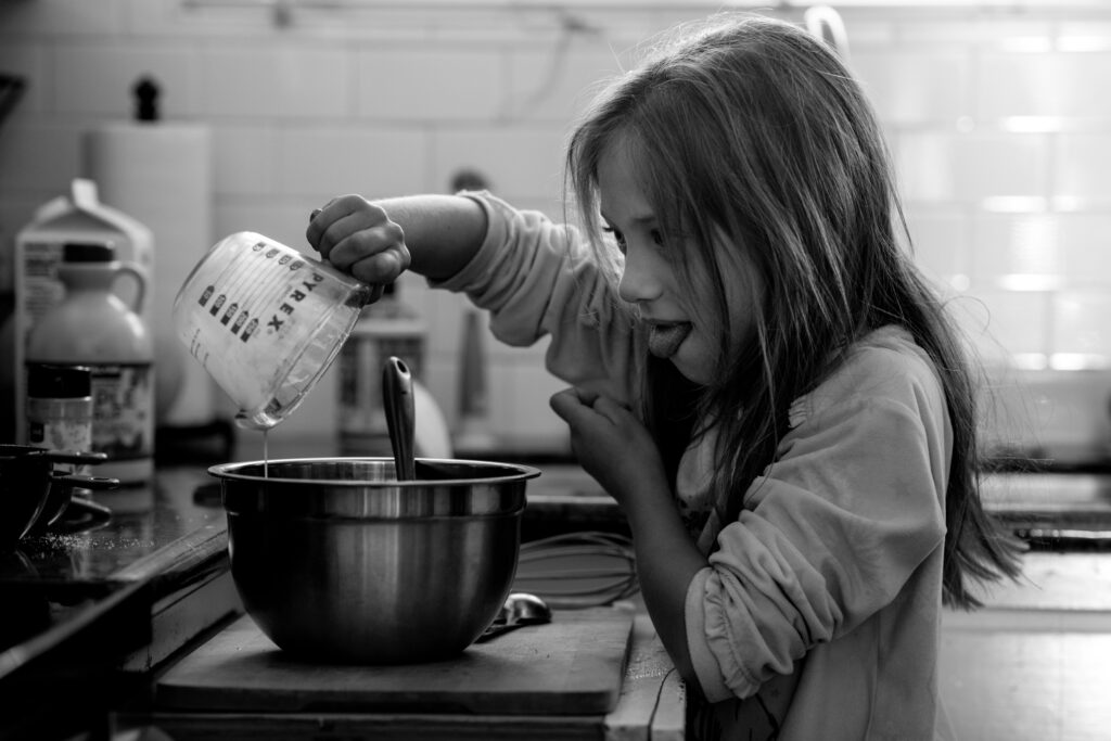 A young girl with messy hair sticks her tongue out as she carefully pours milk into a bowl as she makes pancakes. 