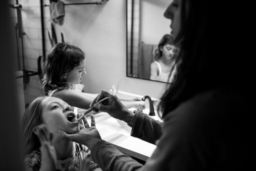 Documentary style family picture of a mom helping her one daughter brush her teeth while her older daughter is in the background getting her toothbrush ready. 