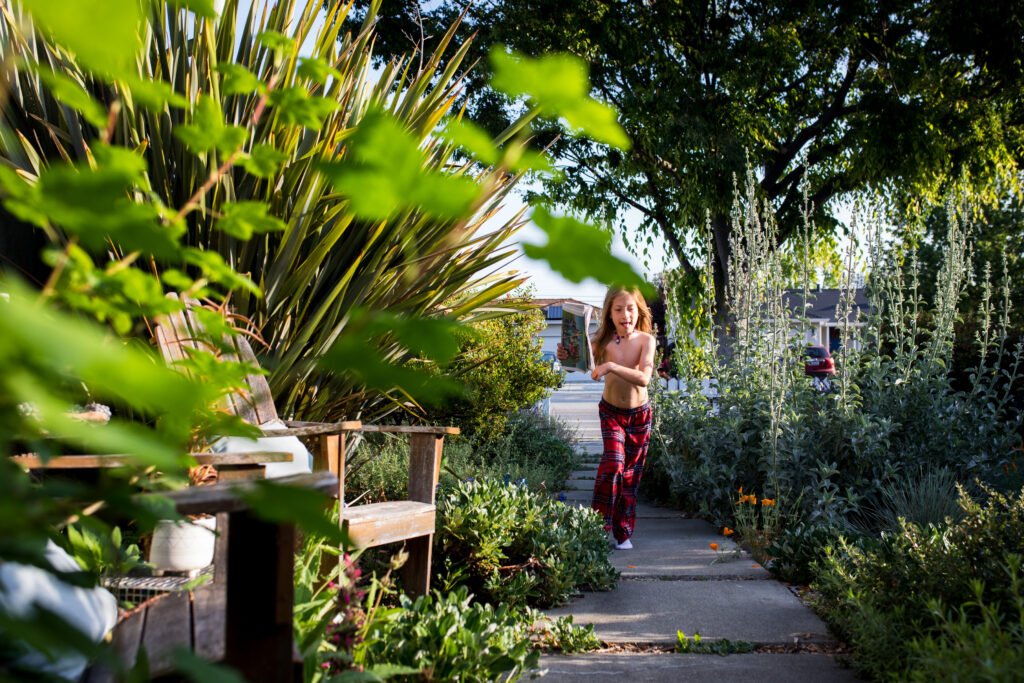 A young girl wearing only red plaid pajama pants runs back towards her front door along the path through the garden holding the newspaper. 