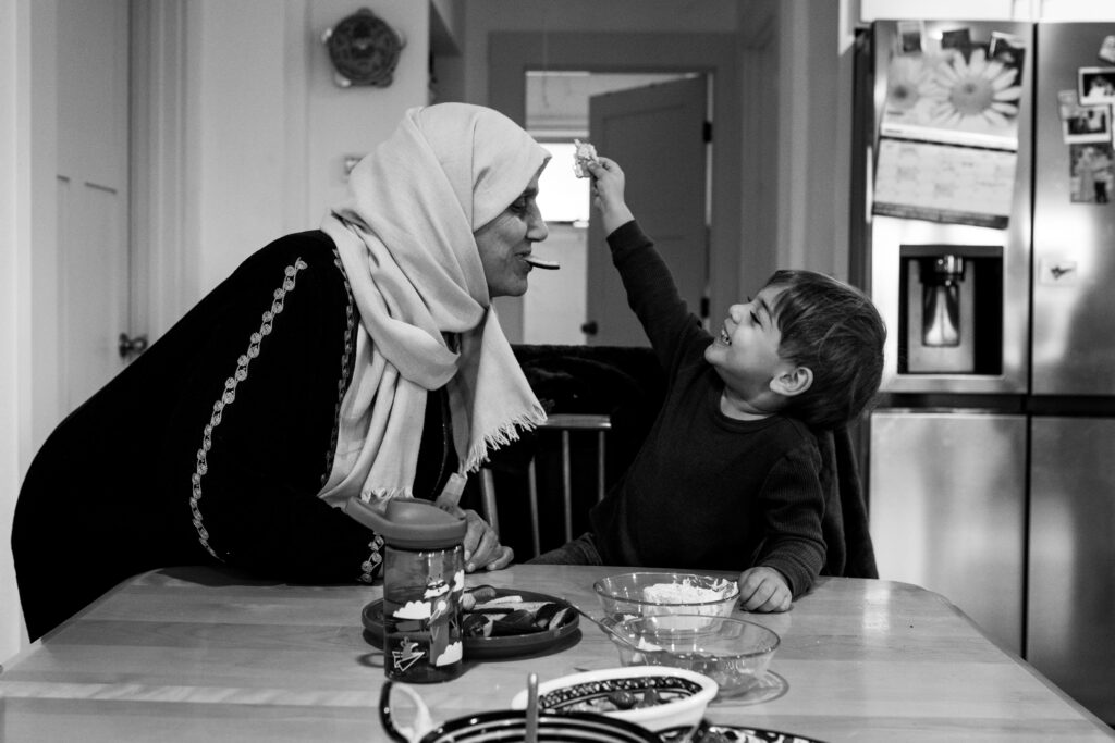 A grandmother in a hijab holds a biscuit in her mouth while making a face at her grandson who is smiling at her and holding another biscuit up to her. 