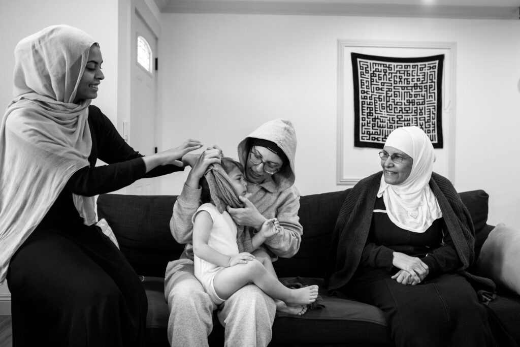 A young boy sits on his mom's lap while she gets him dressed. She is sitting on the couch with her sister in a hijab helps and her mother wearing a hijab looks on. 