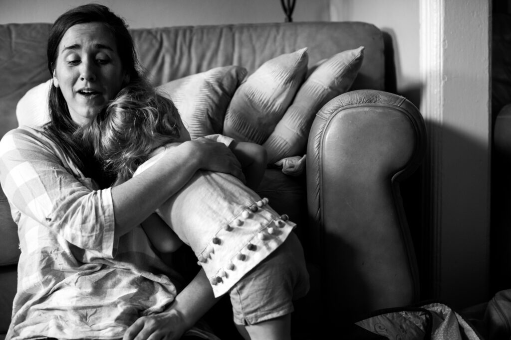The first image in a two image series. Black and white documentary style family photograph of a mom sitting on the floor in front of the couch embracing her toddler after a fall. 
