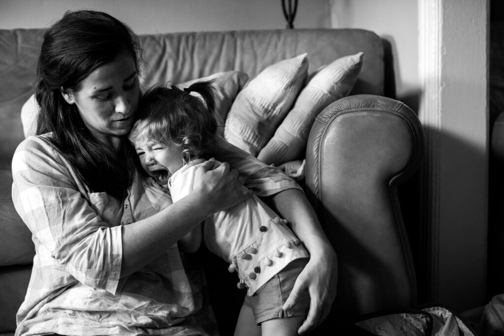 A mom sitting on the floor in front of the couch holds and comforts her screaming toddler. 