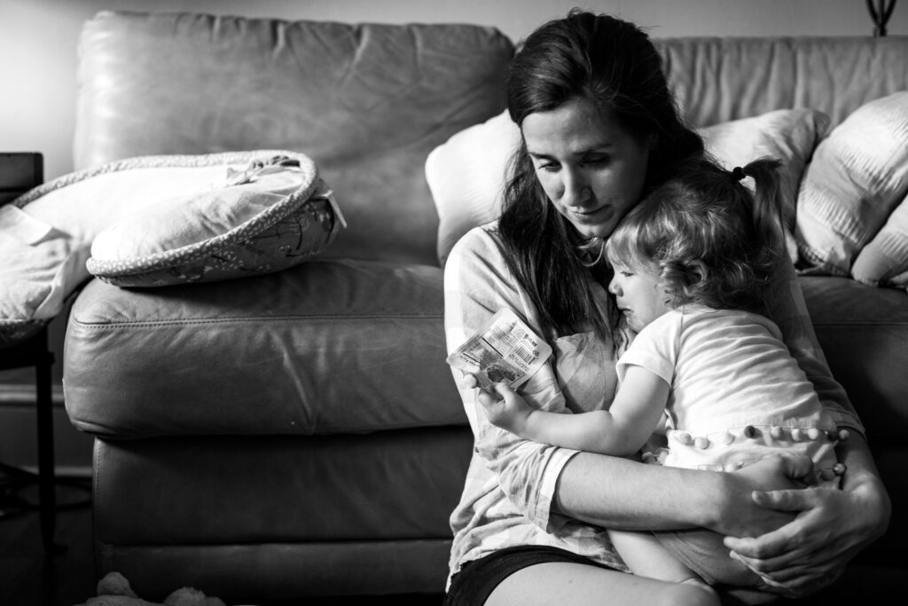 The second image in a two image series. Black and white documentary style family photograph of a mom sitting on the floor in front of the couch embracing her toddler after a fall. The little girl has a very sad face with a full pout and is holding an applesauce pouch.