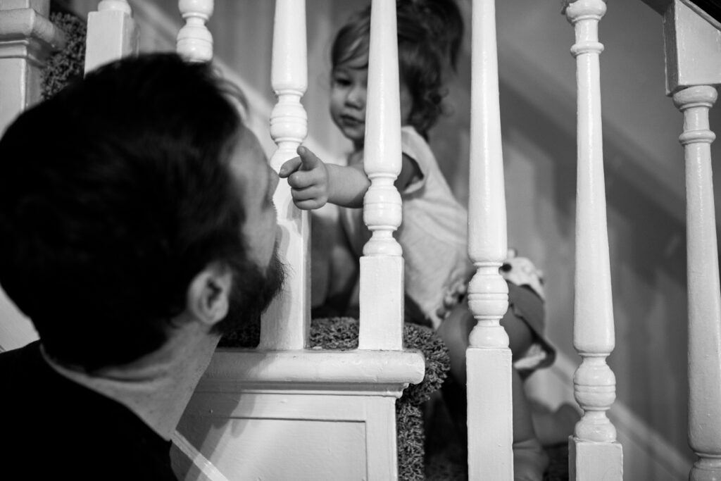 A toddler girl reaches through the railing as she crawls up the stairs to touch her dad on the nose. 