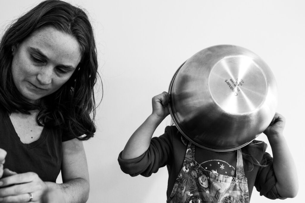 Documentary family photograph in black and white of a mom on the left side of the image looking down at her hands while her toddler on the right has a metal bowl held in front of her face while wearing an animal print apron. 