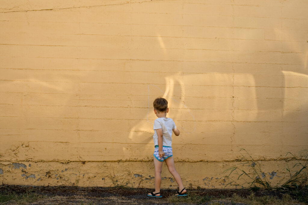 Center weighted image of a little boy wearing a tee shirt and underpants picking a wedgie in front of a yellow brick wall. 