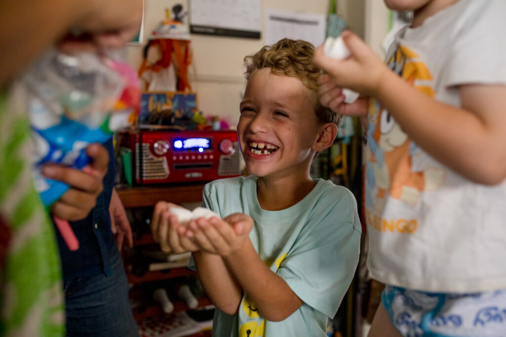 A boy smiles huge with gaps between his teeth as he holding marshmallows in his hand.