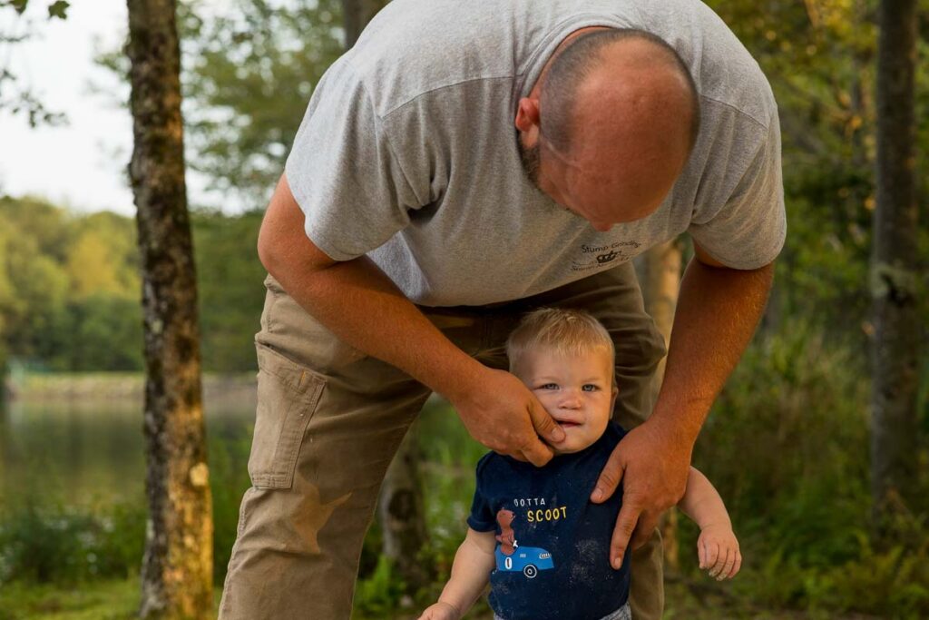 Dad hunches over his toddler boy on the lakeshore, using his thumb to wipe off the boys mouth. 