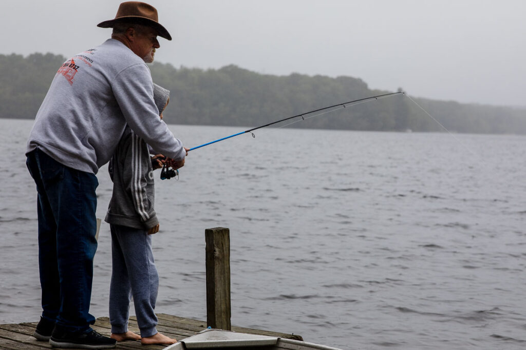 A boy in a sweatshirt with his hood up holding a fishing pole is helped by his grandfather in a cowboy hat as they fish together at the end of a wooden dock on a lake. 