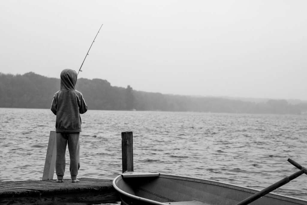 Black and white image of a young boy wearing a sweatshirt with his hood up standing at the end of dock with a fishing pole, fishing in the drizzling rain. 