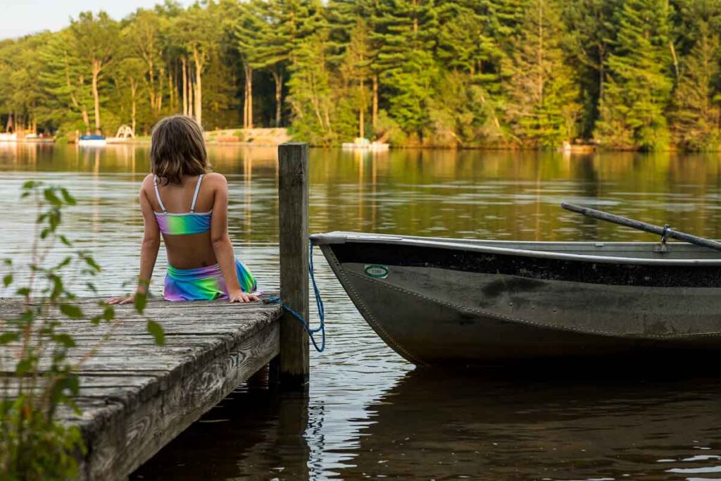A girl in a rainbow neon bathing suit sits on the edge of a small dock on a lake with her feet in the water and leaning back onto her hands. A rowboat floats on the water next to her. 