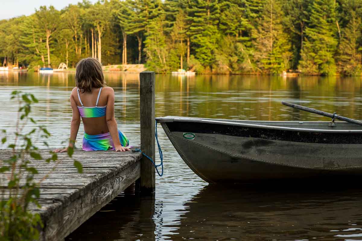 A girl in a rainbow neon bathing suit sits on the edge of a small dock on a lake with her feet in the water and leaning back onto her hands. A rowboat floats on the water next to her.