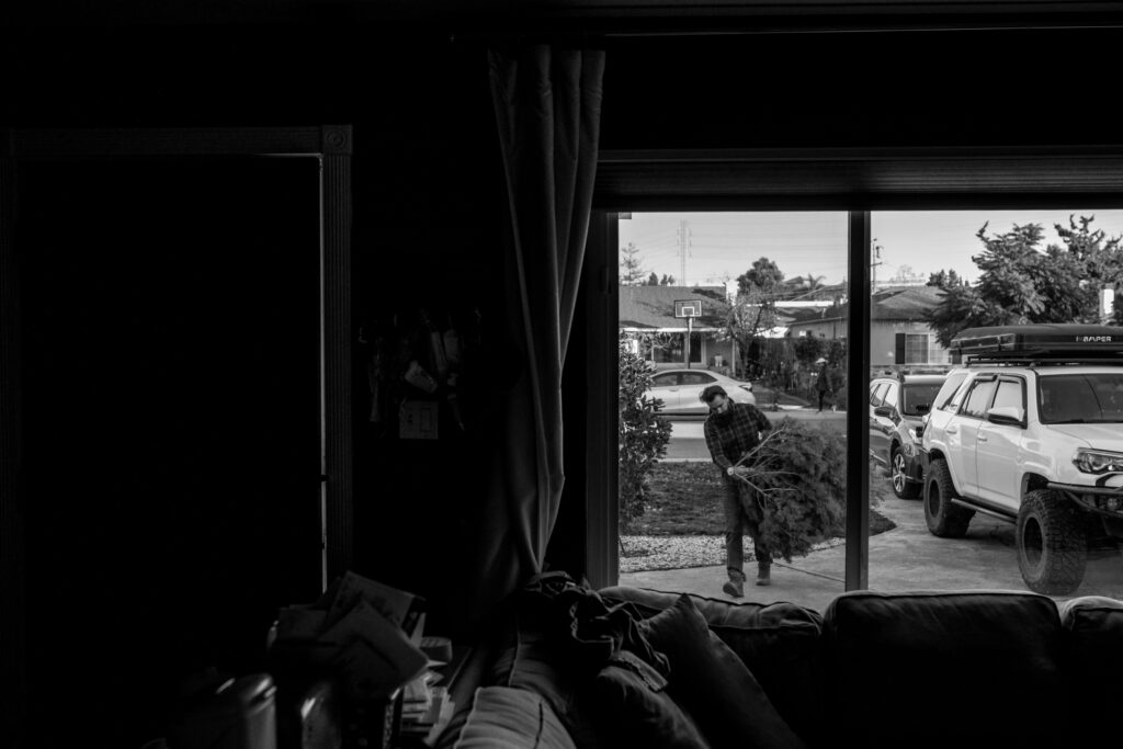 A man outside carries a Christmas tree into the house, photographed from inside the house through the window.